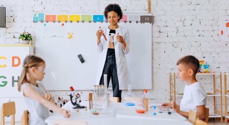 Fotografía de una profesora en una sala de clases, con tubos de ensayo y una pizarra con letras detrás de ella. Dos estudiantes están sentados en una mesa, mezclando líquidos en un tubo de ensayo. La fotografía representa un tipo de docente que se puede ser.