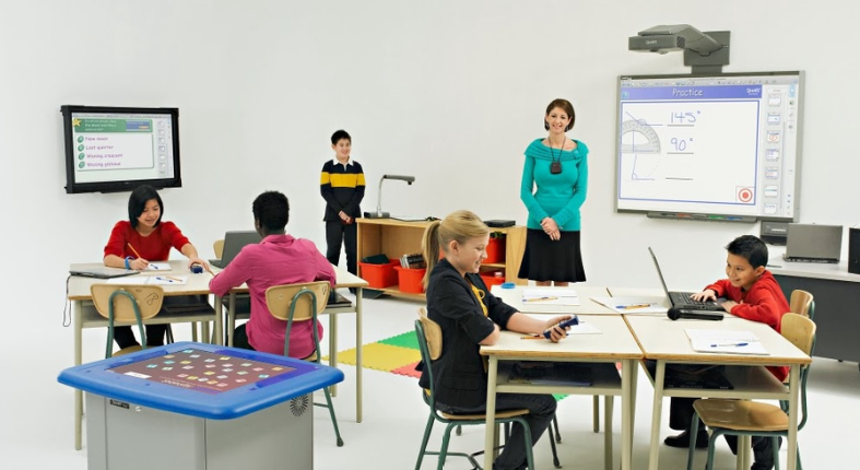 Fotografía de un profesora en una sala de clases con estudiantes interactuando con herramientas tecnológicas, mientras un compañero expone sobre un tema relacionado a la clase de matemáticas. En la pizarra interactiva se puede ver un ejercicio de ángulos. La fotografía representa un tipo de docencia colaborativa y participativa. Imagen de lampadia