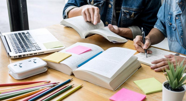 Se observan las manos de dos personas en una mesa, tomando a puntes en una libreta, revisando un libro, hay un computador, post it de notas y lápices de colores. La escena indica que están informándose sobre un tema.