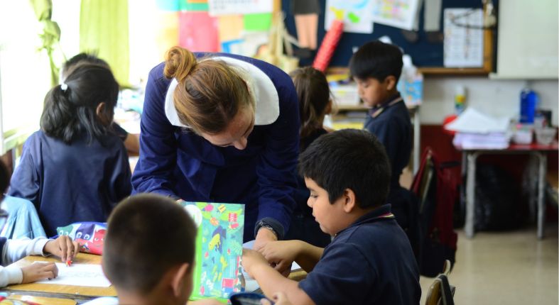 Fotografía de una profesora en una sala de clases, monitoreando el trabajo de sus estudiantes que están dispuestos en grupos. La fotografía representa un tipo de docente que se puede ser.