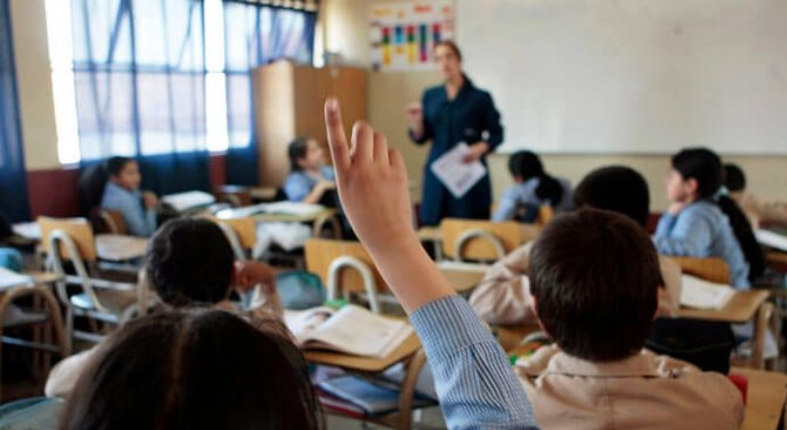 Sala de clases con zoom a una estudiante que levanta su mano para pedir la palabra. La profesora y el resto de sus compañeros y compañeras están fuera foco.