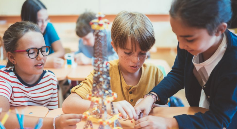 Fotografía de tres estudiantes trabajando en equipo para la elaboración de un tipo de torre. Detrás de ellos se observan otros estudiantes en los mismo.