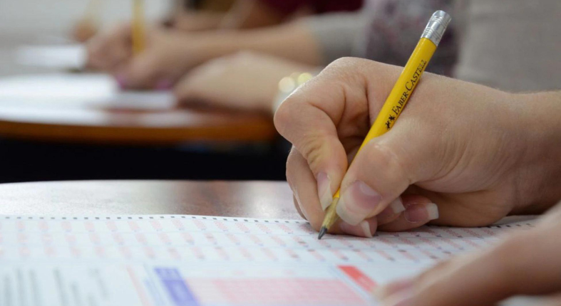 En la imagen se observa en primer plano la mano de un estudiante rindiendo una prueba de selección. En el fondo desenfocado se aprecian otros estudiantes haciendo lo mismo.