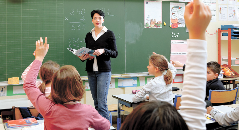 Fotografía de una profesora en una sala de clases, mirando a sus estudiantes que se ven levantando la mano para participar. La fotografía representa un tipo de docente que se puede ser.